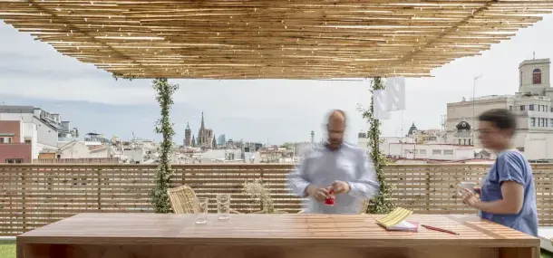 Una mesa de madera sostiene vasos vacíos mientras dos personas se mueven rápidamente bajo un techo de bambú en una terraza, con edificios urbanos al fondo.