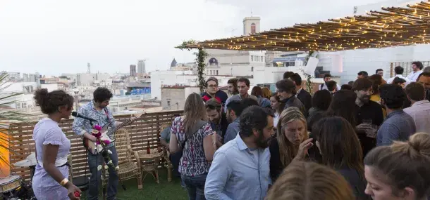 Varias personas socializan en una terraza decorada con luces, con vistas a la ciudad en el fondo.