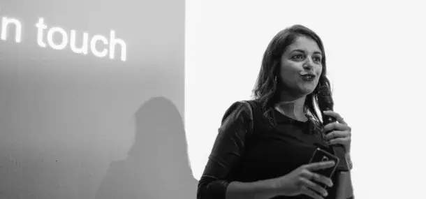 Una mujer sosteniendo un micrófono está hablando. Lleva un teléfono en otra mano. En el fondo, hay una pantalla con texto parcial: "in touch."