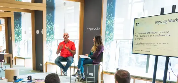 Hombre y mujer sentados conversan en una sala moderna con ventanas; pantalla muestra: "Inspiring Work, Claves para una cultura corporativa feliz y su impacto en el crecimiento de la empresa, Con Jaume Gurt, uno de los influencers más destacados de nuestro país y antiguo director de InfoJobs, Apúntate en: utopicus.es/agenda".