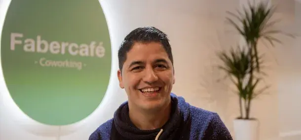 Un hombre sonríe en un entorno de oficina. Al fondo, un cartel verde dice "Fabercafé - Coworking", rodeado de plantas en macetas.