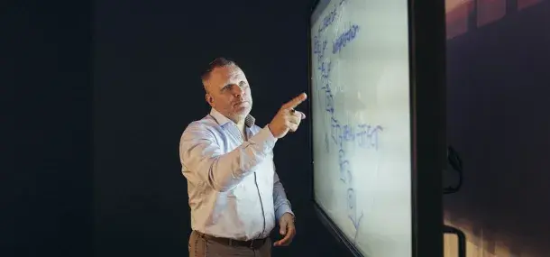 Un hombre señala una pizarra interactiva con ecuaciones en un aula oscura, concentrado en la explicación. No hay texto visible específico.