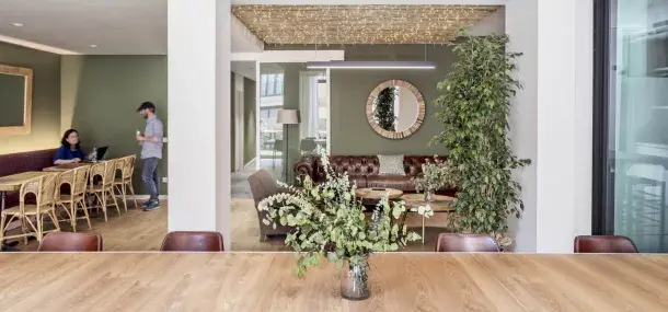Una mesa de madera sostiene un florero con hojas verdes, ubicada en una sala decorada con sofás, espejos y plantas. Al fondo, personas conversan junto a mesas y sillas de mimbre. Ambiente moderno y acogedor.