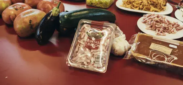 Sobre una mesa roja, un paquete de "cintas de bacon" está rodeado de calabacines, berenjenas, tomates, ajo y platos con alimentos variados, creando un ambiente culinario.