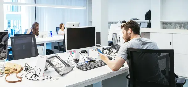 Un hombre trabaja en una oficina, mirando papel y computadora en un escritorio desordenado, mientras otras personas están sentadas al fondo.