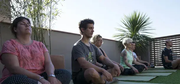 Personas meditando sentadas sobre colchonetas verdes, en una terraza rodeada de plantas y con cielo despejado.