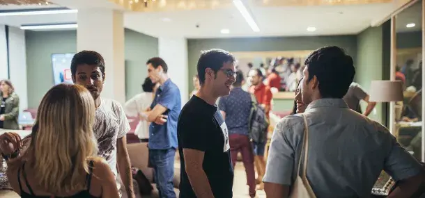 Varias personas conversando en un salón iluminado, rodeados de decoración moderna y otros asistentes al fondo.