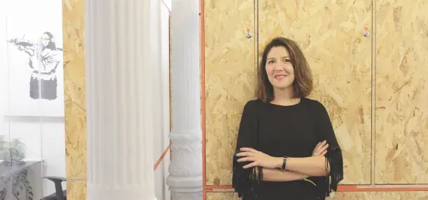 Una persona de pie con brazos cruzados, sonriendo, está frente a paredes de madera y una columna clásica en una oficina moderna. A la izquierda, hay una imagen en blanco y negro enmarcada.