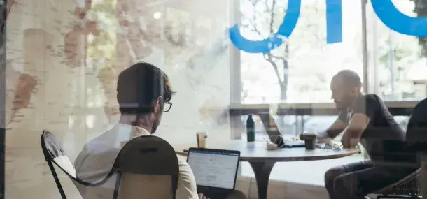 Personas trabajando en laptops, sentadas alrededor de una mesa redonda, dentro de una oficina con ventanas grandes. Letras azules parcialmente visibles en el vidrio.