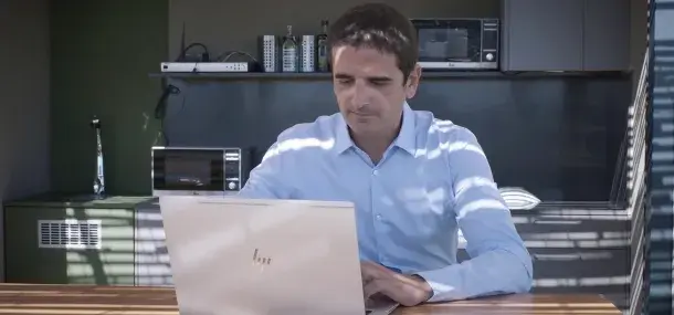 Un hombre usando una computadora portátil está sentado en una cocina, iluminado por luz que entra a través de persianas. Alrededor, se ven electrodomésticos como microondas y estantes con botellas.
