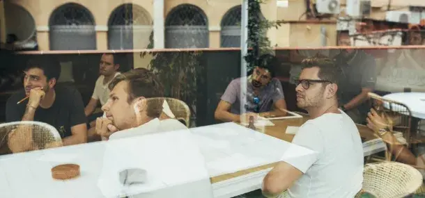 Varias personas están sentadas en una mesa, mirando atentamente en la misma dirección, en un ambiente interior con ventanas grandes.
