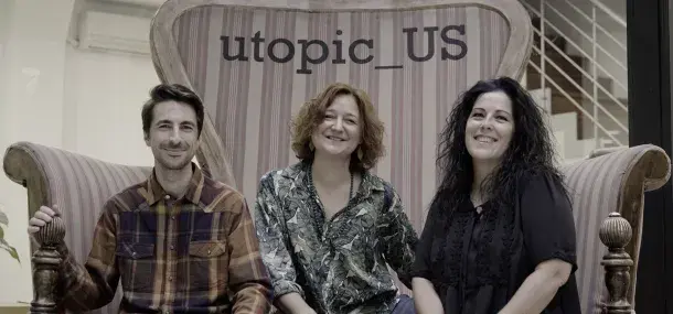 Tres personas sentadas en un sofá grande, sonriendo; detrás, el texto "utopic_US". Ambiente interior con escaleras al fondo.