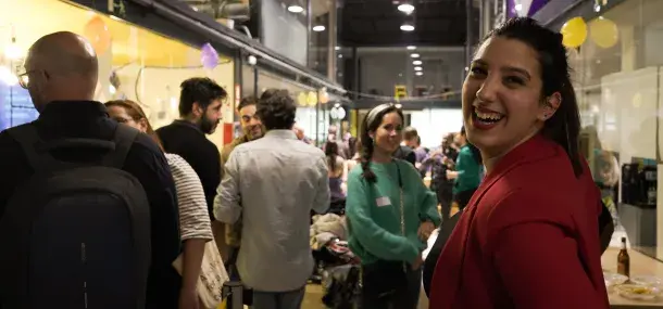 Persona sonriendo, vistiendo chaqueta roja, en un evento social concurrido con personas conversando en un espacio cerrado decorado con globos.