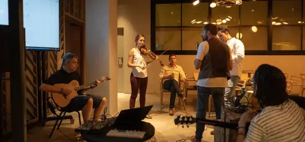 Un grupo de personas toca instrumentos musicales, como la guitarra y el violín, en una sala iluminada. Algunos están sentados, otros de pie, rodeados de equipos electrónicos.