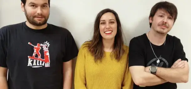 Tres personas de pie, sonriendo frente a una pared blanca. Una lleva camiseta negra con texto: "Rock de aki Kalla".