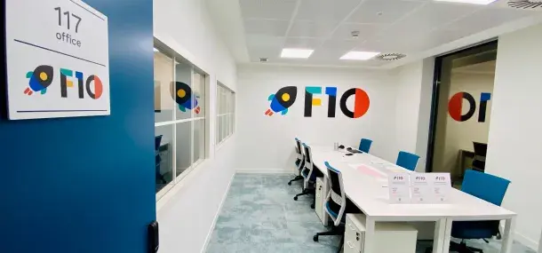Una sala de reuniones moderna con seis sillas azules alrededor de una mesa blanca. En la pared, el texto "F10" y un dibujo de cohete. La puerta tiene un cartel: "117 office".