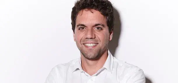 Un hombre sonriente lleva una camisa blanca, de brazos cruzados, sobre un fondo blanco liso.