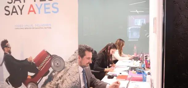 Varias personas trabajando en un escritorio en una oficina moderna. Cartel publicitario dice "SAY YES SAY AYES" y "ADDED VALUE, DELIVERED".