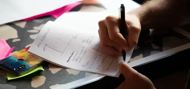 Una mano escribe con un bolígrafo en un papel con gráficos y texto en un escritorio con un teléfono colorido bajo papeles.