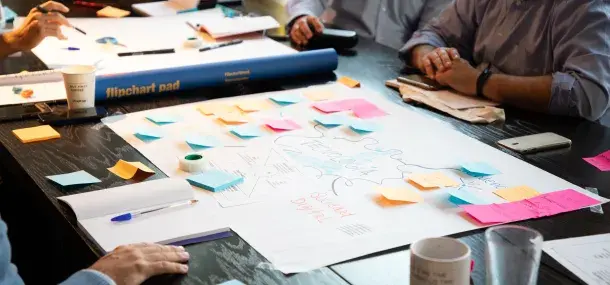 Una mesa cubierta de notas adhesivas y papeles. Personas escriben y discuten. Ambiente de reunión colaborativa.