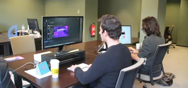 Un hombre trabaja en una computadora con un editor de texto abierto, en una oficina compartida. Sobre la mesa, un cartel dice "DO NOT DISTURB" y una taza "BLONDE ROAST".