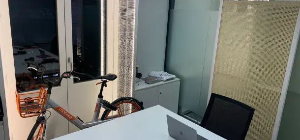 Una bicicleta estática se encuentra al lado de una mesa blanca con una laptop abierta en una oficina moderna con paredes de vidrio y muebles minimalistas.