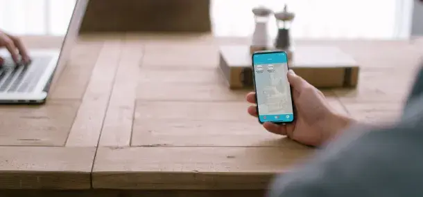 Un teléfono móvil sostenido por una mano muestra una pantalla azul con iconos. En una mesa de madera, al fondo, hay una computadora portátil y utensilios de cocina.