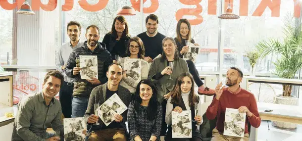 Un grupo de personas sostiene revistas sonrientes en una cafetería luminosa y moderna. Al fondo, un cartel rojo parcialmente visible.