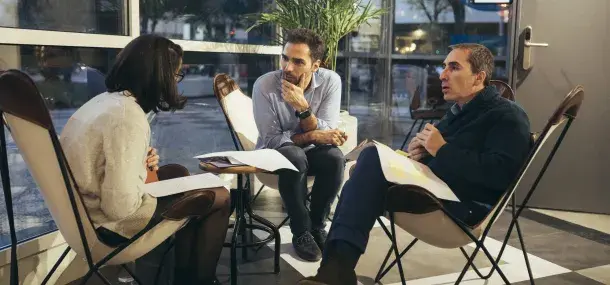 Tres personas sentadas en sillas, discutiendo documentos, en un ambiente interior junto a una ventana grande.