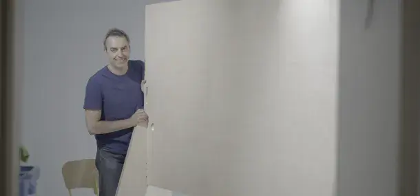 Un hombre, sonriendo, sostiene una puerta grande de madera en una habitación clara con una silla al fondo.
