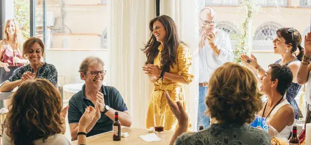 Una mujer de pie sonríe mientras varias personas aplauden a su alrededor en una reunión social informal. Están en una sala iluminada junto a una ventana grande.