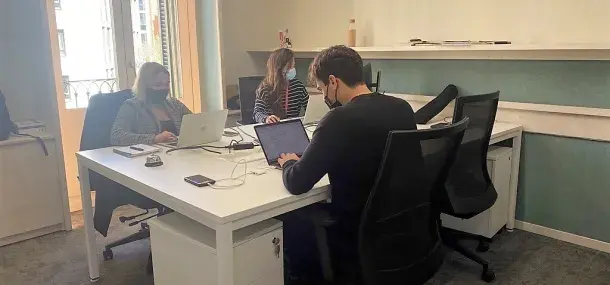 Tres personas trabajan en laptops, sentadas alrededor de una mesa blanca en una oficina iluminada por luz natural.