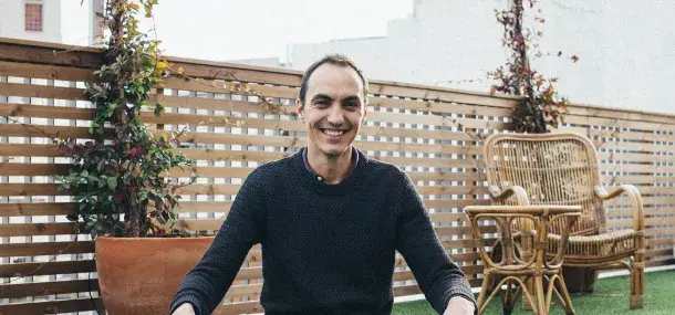 Un hombre sonriente está sentado en una terraza. A su alrededor, hay plantas y sillas de mimbre.