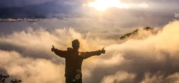 Una persona con brazos extendidos y pulgares levantados mira hacia un amanecer entre nubes, sobre una montaña.