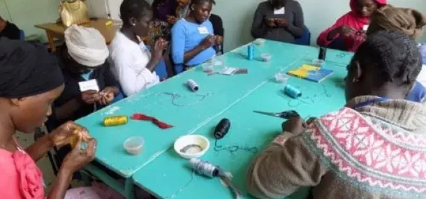 Un grupo de personas realiza manualidades con hilos en una mesa azul; hay bobinas y herramientas dispersas alrededor. Están concentrados y colaborando en un espacio interior.