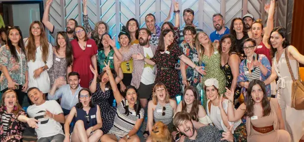 Un grupo de personas sonríe y hace gestos alegres. Están en un ambiente festivo con decoración luminosa. Un perro se encuentra entre ellos en el frente.