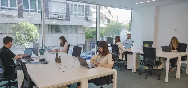 Personas trabajando en laptops. Están sentadas en escritorios blancos en una oficina iluminada con una gran ventana que muestra un edificio y árboles afuera.