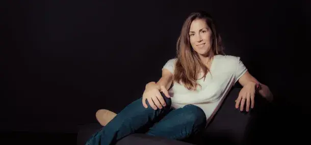 Una mujer está sentada relajadamente en un sillón negro, con un fondo oscuro, vistiendo una camiseta blanca y jeans.