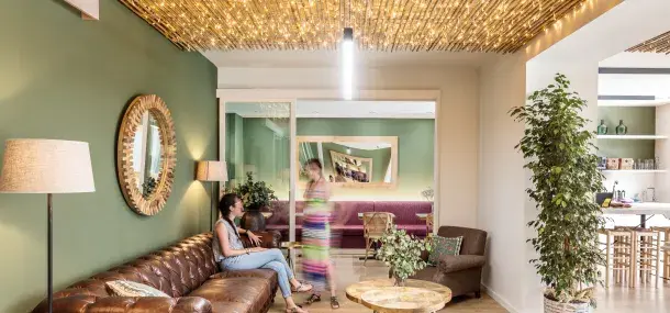 Una mujer está sentada en un sofá de cuero marrón, hablando con otra que pasa caminando. La habitación tiene paredes verdes, un techo con luces cálidas, espejos y plantas decorativas.