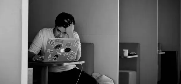 Persona concentrada usando una laptop llena de pegatinas, en una sala de estar, apoyando su cabeza con una mano y sosteniendo la otra en el teclado.