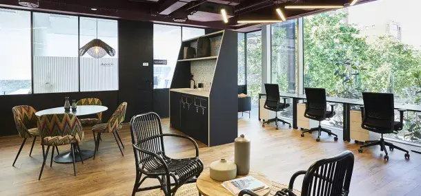 Espacio de oficina moderno; mesa y sillas en el centro, escritorios al fondo junto a ventanas grandes, con luz natural y vista a árboles. Lámparas colgantes iluminan el área.
