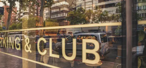 Reflejo de la calle en una ventana con letras grandes que dicen "WORKING & CLUB", rodeado de edificios y árboles.