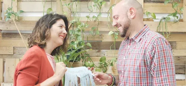 Una mujer y un hombre sonríen sosteniendo un papel con texto ilegible. Están en un entorno con palés y plantas verdes colgantes.