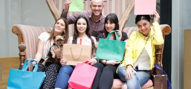 Varias personas sonrientes sostienen bolsas de compras de colores mientras están sentadas en un sofá estilizado en un entorno interior. Una persona sostiene un perro pequeño.