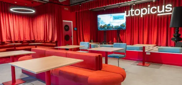 Sofás rojos y mesas vacías en un salón con cortinas rojas, pantalla grande y el texto "utopicus".