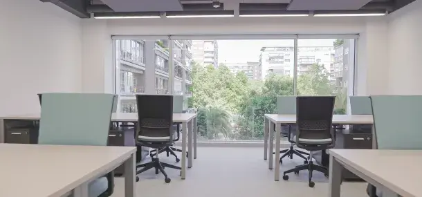 Escritorios vacíos con sillas giratorias están frente a una ventana grande que muestra edificios y árboles afuera.