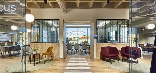 Una moderna sala de reuniones con mesas y sillas, iluminación colgante, y puertas abiertas hacia el exterior urbano. La pared de cristal muestra reflejos, añadiendo elegancia al ambiente.
