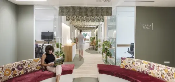 Una mujer está sentada trabajando en un portátil en un sofá curvo con cojines florales. Al fondo, un hombre camina por un pasillo decorado con plantas y lámparas. En la pared, se lee "ICARIA PLAZA".