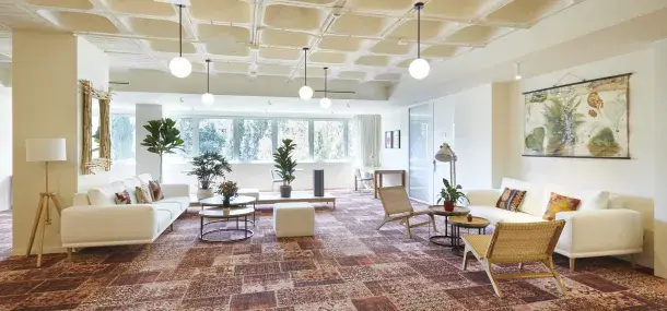 Sofás blancos organizados con mesas de centro y plantas, en una sala iluminada por grandes ventanales, con lámparas colgantes y paredes decoradas.
