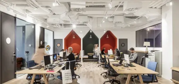 Varias personas trabajan en sus computadoras en una oficina moderna, con cabinas de privacidad coloridas y decoración minimalista.